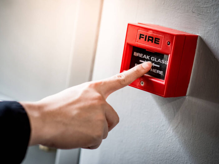 fire risk assessment – Male hand pointing at red fire alarm switch on concrete wall in office building.